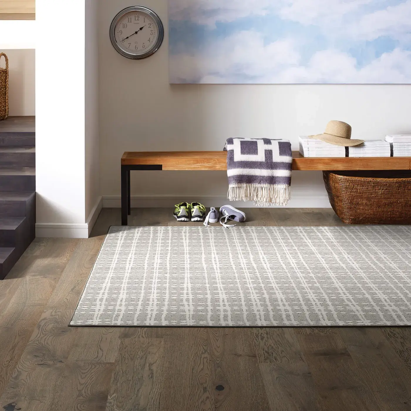 CastlewoodOak-9U899-00508-Armory-PureEnvy-E9361-00100-Snowfall copy A light-colored Shaw Floors area rug with a modern, textured striped pattern on a dark wood floor in an entryway with a wooden bench, clock, and staircase leading down.