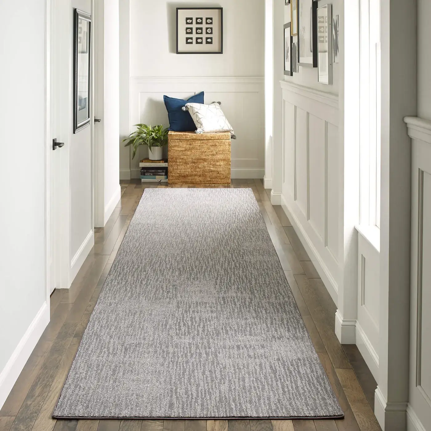 DynamicVision-HDE96-00501-Houndstooth-Hallway-2-V A long, grey Shaw Floors area rug with a subtle textured pattern runs down a bright white hallway with dark wood floors, leading to a small bench with pillows and framed artwork.