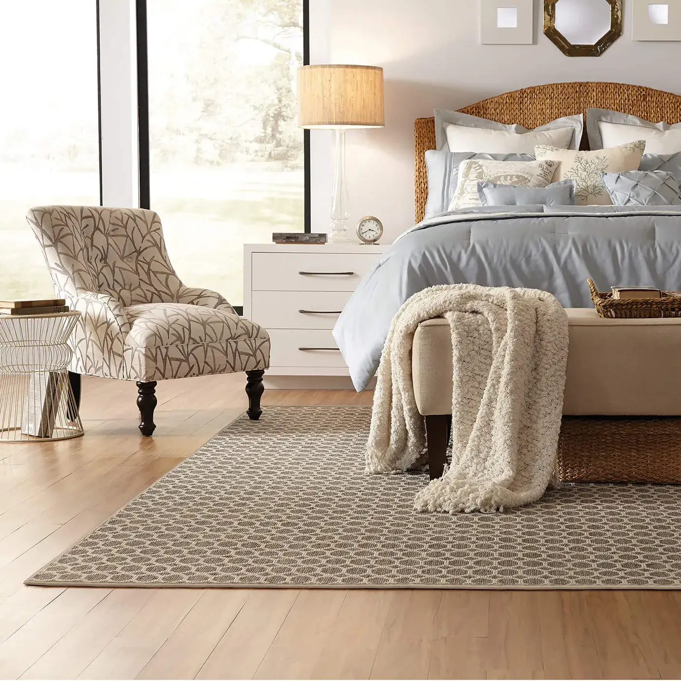 TRACERY-Z6878-RUG-H copy A patterned Anderson Tuftex area rug in shades of brown and beige with a circular design on a light wood floor in a bedroom with a rattan headboard bed and a patterned accent chair.